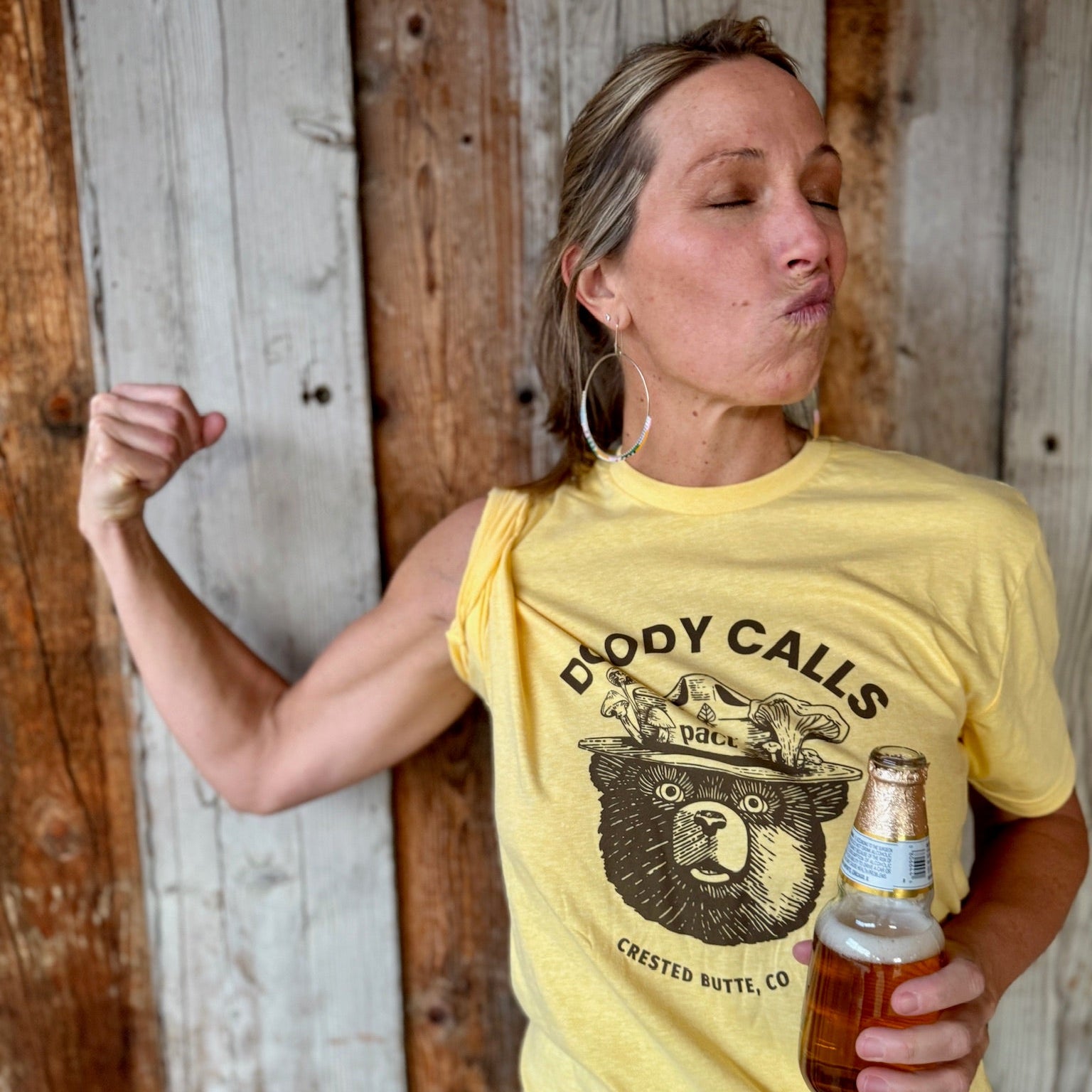 Woman wearing a yellow t-shirt with a bear graphic and text, holding a beer bottle against a wooden background.
